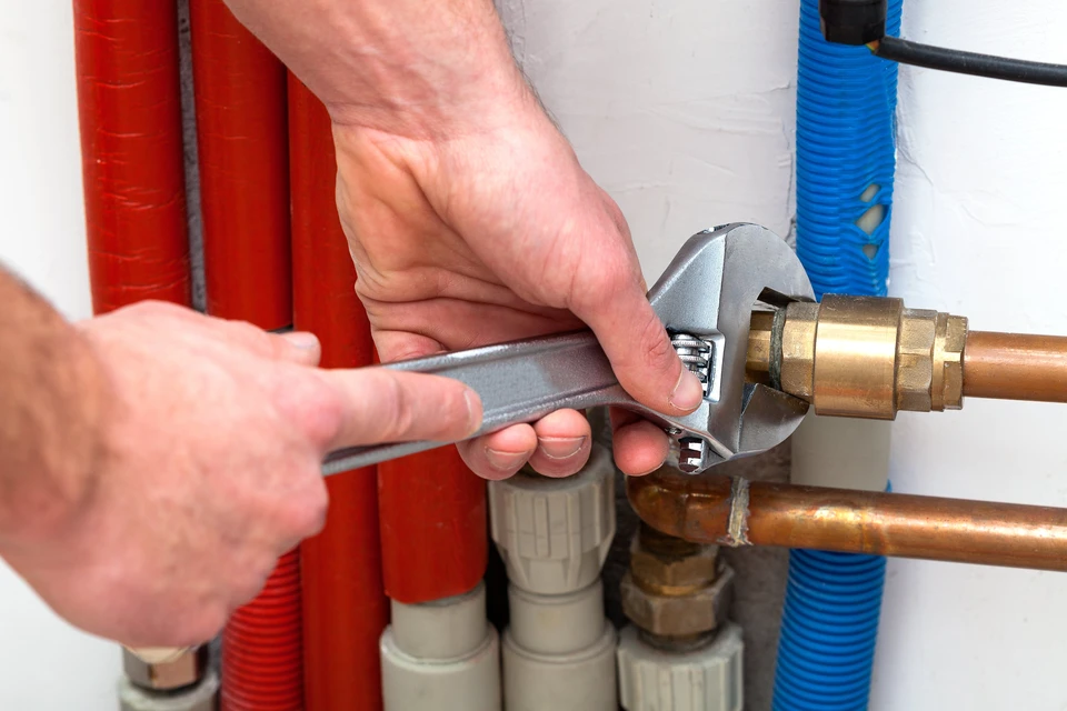 Homme en chemise à carreaux effectuant des travaux de plomberie sur un radiateur blanc dans un intérieur résidentiel lumineux près d'une fenêtre